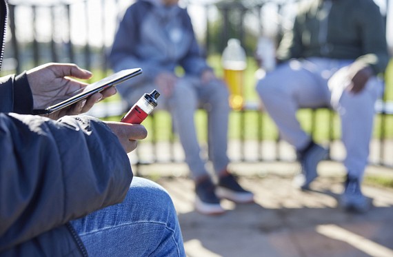 Three teenagers sitting in a park with a vape and phone in their hands.
