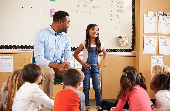 young child speaking in front of a class next to the teacher